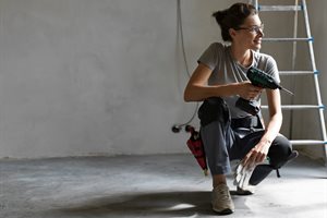 Female with glasses crouching. She is holding a drill and work gloves. There is a ladder and cable behind her.