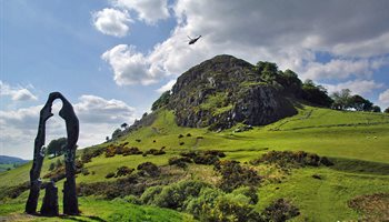 Silhouette of a tall person which is the Spirit of Scotland sculpture, Loudoun Hill in the distance and a helicopter flying past