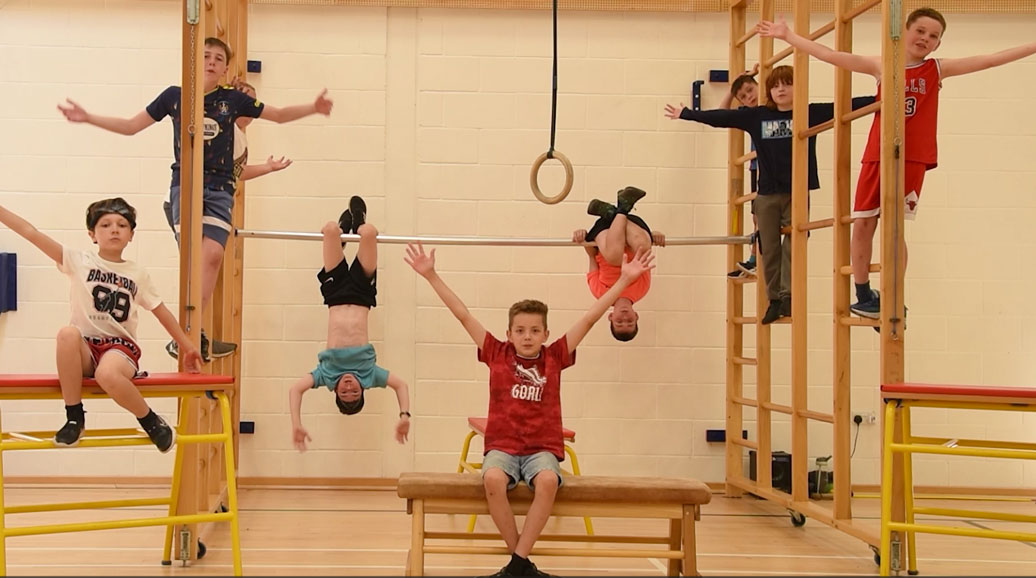 young boys in a school gym hall, sitting on activity benches, standing on climbing frames and hanging upside down on a metal pole