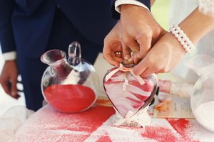 Two sets of hands sealing a clear glass heart container with red and white sand