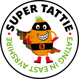 A smiling orange super tattie with green spikey hair, black eye mask, black belt with S on it, red gloves and boots and a tartan cape. Super Tattie is the Mascot for primary school meals in East Ayrshire. The words around Super Tattie say "Super Tattie, E