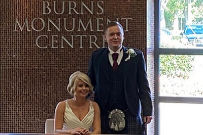 Bride sitting and groom standing at a table, smiling, at the Burns Monument Centre