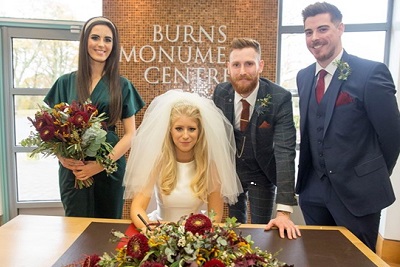Bride sitting and groom standing at a table, smiling, guests beside them, at the Burns Monument Centre