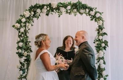 Bride and groom laughing, standing under a white flower arch and the registrar laughing