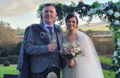 Bride and groom smiling, white flower arch and countryside in the background