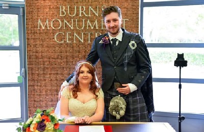 Bride sitting and groom standing at a table, smiling, at the Burns Monument Centre