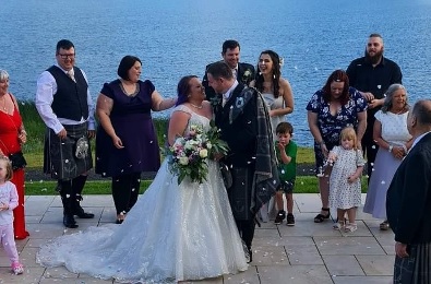 Bride and groom looking into each other eyes smiling, water in the background and guests around them