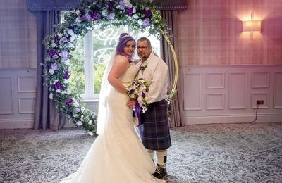 Bride and groom smiling, purple and white flower arch in the background beside a large window