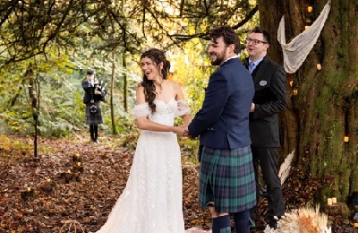Bride and groom holding hands smiling, standing beside a tree getting married, piper in the background